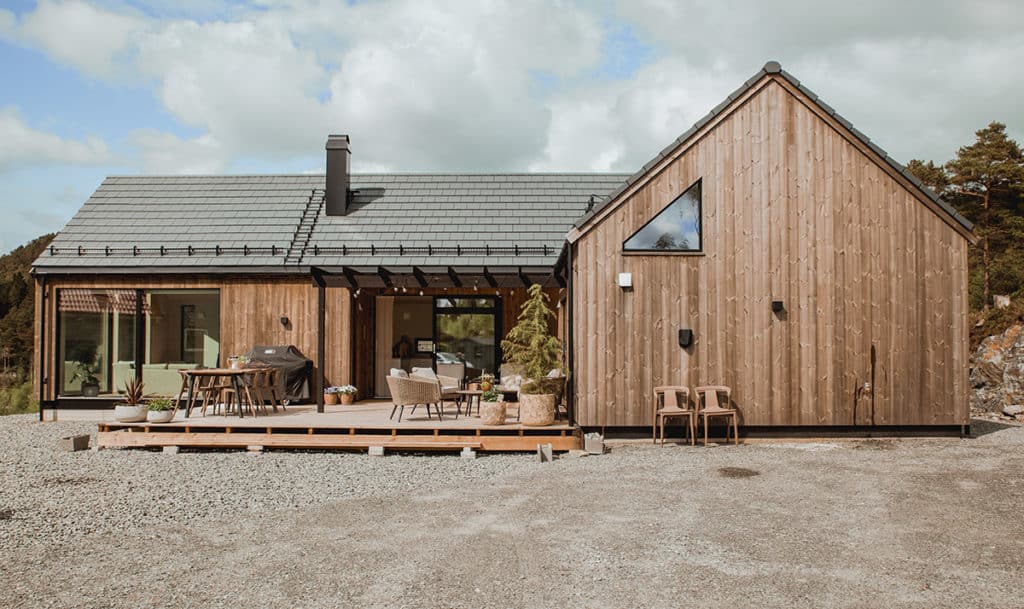 Trekledd hytte med saltak, store vinduer og møblert terrasse